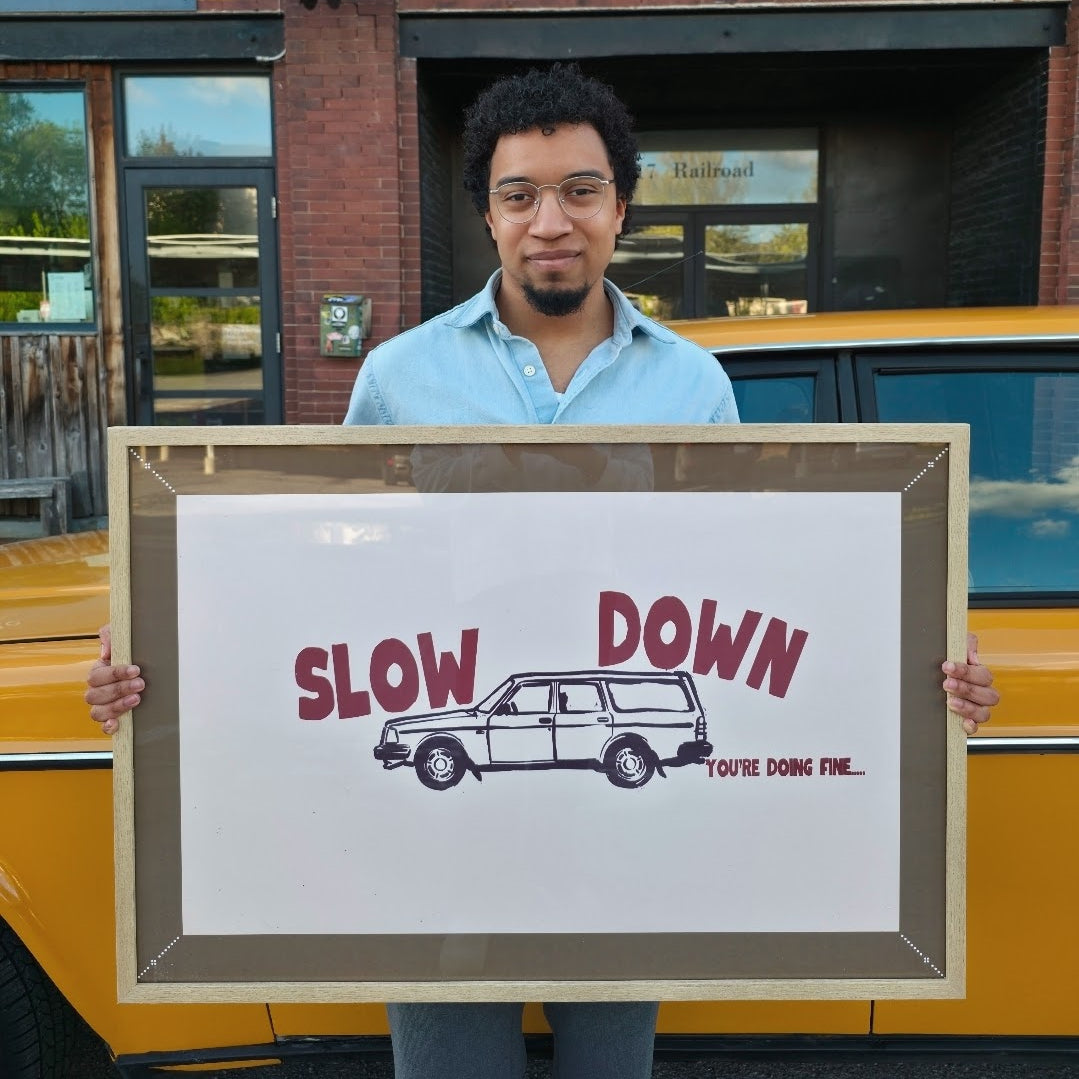 Person holding a framed picture with 'Slow Down' text and car illustration in front of a building.